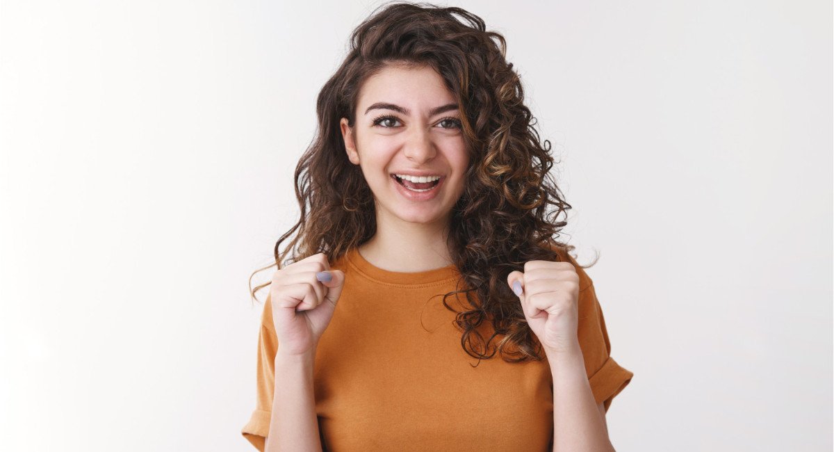 A person with curly hair is raising their fists in a playful and energetic pose, wearing a casual brown shirt against a light background.