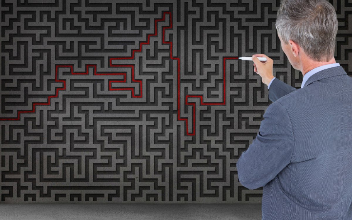 A businessman in a suit points at a complex black maze on a wall, highlighting a pathway traced in red marker.