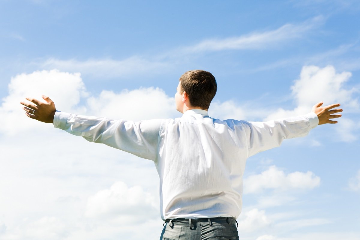 A man in a white shirt stands outdoors, arms outstretched, embracing the blue sky and fluffy clouds above him.
